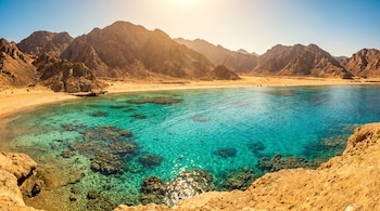 Sol brillante sobre cielo azul, montañas desérticas, playa de arena, mar turquesa con arrecifes de coral y costa rocosa