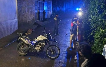 Una motocicleta sobre una calle de noche, con una persona en el suelo, agentes policiales y vecinos observando bajo luces azules