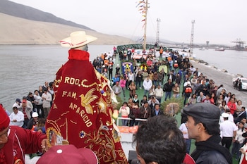 Vista trasera de una estatua con capa roja bordada y sombrero de paja en la cubierta de un barco, rodeada de una multitud de personas. Al fondo, una bahía y montañas