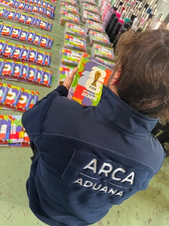 Vista superior de una persona con chaqueta azul con el logo ARCA Aduana, sosteniendo un álbum FIFA World Cup 2026. Hay paquetes de figuritas en el fondo
