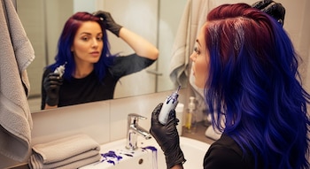 Mujer con guantes negros tiñe su cabello de colores azul y rojo-púrpura frente a un espejo en el baño, sosteniendo un tubo de tinte.