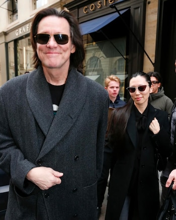 Jim Carrey con un abrigo gris de espiga y gafas de sol camina por la calle junto a Min Ah