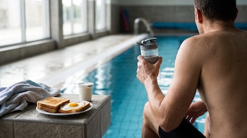 Un hombre de espaldas sostiene una botella junto a una piscina, al lado de un plato con tostadas y un huevo frito, un yogur y una toalla.