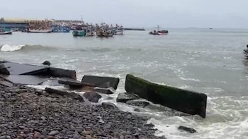 Mar con oleaje y una orilla rocosa con fragmentos de concreto. En el fondo, varios barcos pesqueros permanecen en un puerto bajo un cielo gris