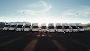 Once camiones blancos modelo Tesla Semi aparcados en fila sobre un asfaltado al atardecer con logotipos de empresas en la parte superior