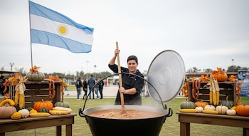 Joven sonriente revuelve guiso en olla gigante al aire libre. Mesas rústicas con calabazas, maíz y chorizos a los lados, bandera Argentina ondeando de fondo.