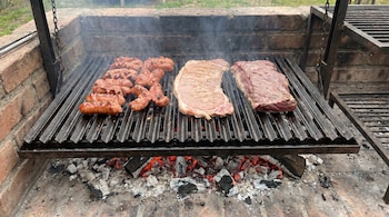 Vista cercana de una parrilla de ladrillos con una rejilla de metal que sostiene chorizos abiertos, matambre de cerdo y un corte de vacío, con brasas rojas visibles debajo y humo.