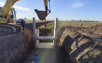Uma escavadeira amarela coloca uma estrutura de concreto pré-moldado em um buraco profundo em uma área rural. Você pode ver as pessoas e a terra escavada