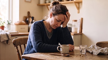 Mujer sentada a una mesa de madera, con la mano izquierda en la frente, una taza beige, un vaso de agua, una cuchara de madera y migas.