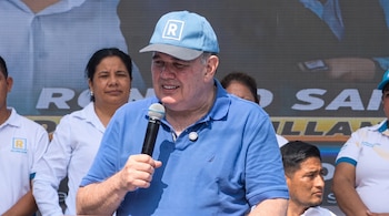 Hombre de mediana edad con gorra azul y polo azul sostiene micrófono. Alrededor, otras personas y una pantalla LED con letras.