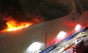 Vista aérea nocturna de un edificio con llamas y humo en el techo, luces rojas y blancas en la pared, y un camión en la calle mojada