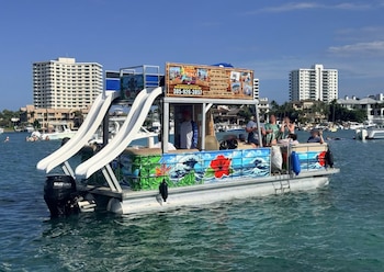 Barco pontón de colores con dos toboganes blancos y un motor Suzuki. Personas a bordo y nadando en aguas claras, con edificios en la costa bajo un cielo azul