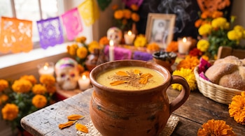 Taza de barro con atole, canela y pétalos, junto a pan de muerto, flores de cempasúchil, veladoras y papel picado en una mesa de madera.