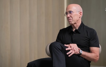 Un hombre calvo con gafas, Jeffrey Katzenberg, viste una polo negra y pantalones oscuros, sentado con las manos entrelazadas y mirando a la izquierda