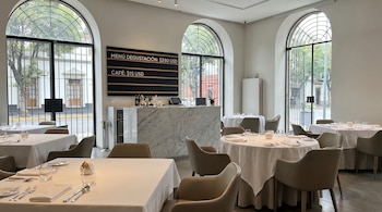Interior de un restaurante de lujo vacío con mesas vestidas de blanco y sillas de cuero. Grandes ventanales arqueados, una barra de mármol y un menú visible.