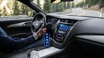 Interior de un automóvil moderno con conductor con guante marrón operando la palanca de cambios automática. Las letras P, R, N, D, L están iluminadas. Al fondo, una carretera de montaña.