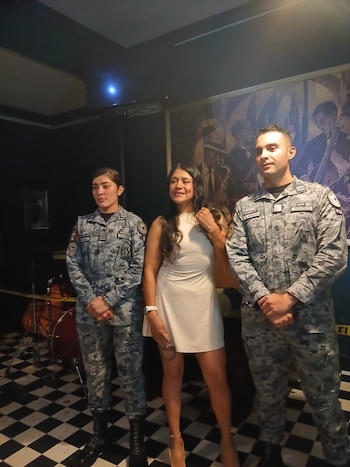 Una mujer vestida de blanco permanece entre dos integrantes de la Guardia Nacional en un recinto con piso de ajedrez
