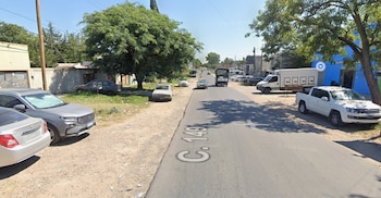 Una calle asfaltada con automóviles, camionetas y camiones estacionados a los lados, rodeada de árboles y edificios bajo un cielo despejado