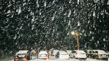 Vista nocturna de una calle bajo una intensa nevada, con vehículos cubiertos de nieve y sus luces traseras encendidas, junto a una farola iluminada