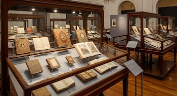 Sala de museo con piso de madera y varias vitrinas de madera y cristal que exhiben numerosos libros antiguos, algunos abiertos mostrando páginas ilustradas.