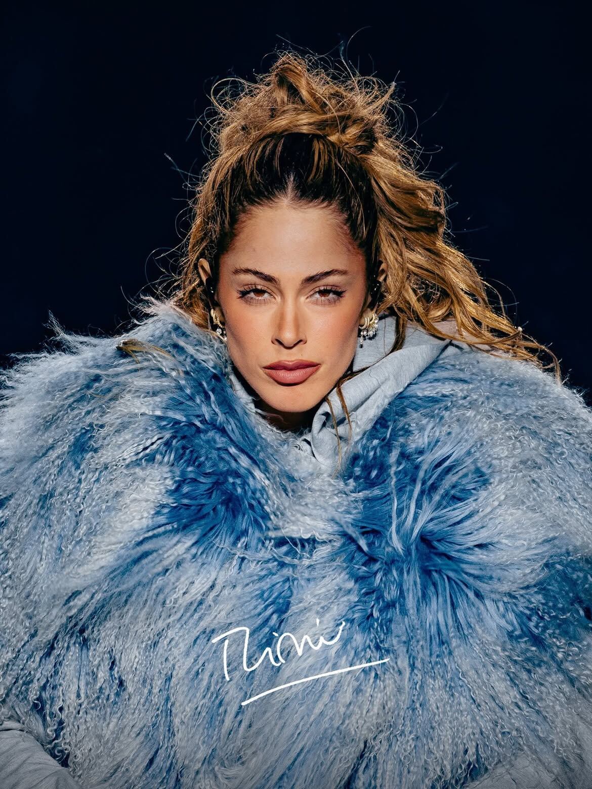 La cantante Tini viste un abrigo afelpado de color celeste sobre un fondo oscuro.