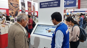 Hombre con saco observa pantalla táctil. Orientador con chaleco azul señala mapa en la pantalla. Mujer con mochila mira la pantalla. Libros y carteles visibles.
