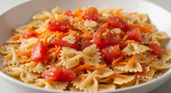 Un plato hondo con fideos moñito, zanahoria rallada, trozos de tomate, rebanadas de ajo, pimienta negra molida y sal.