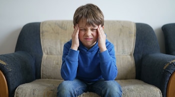 Un niño con sudadera azul y pantalones de jean está sentado en un sofá, con los ojos cerrados y las manos sobre las sienes.