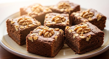 Primer plano de siete brownies cuadrados de avena y cacao sobre un plato artesanal. Cada brownie está decorado con una nuez y trozos, con un fondo luminoso.