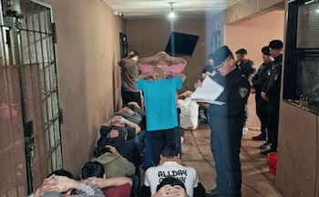Varios detenidos sentados y de pie con las manos sobre la cabeza en un pasillo, mientras oficiales de policía los supervisan
