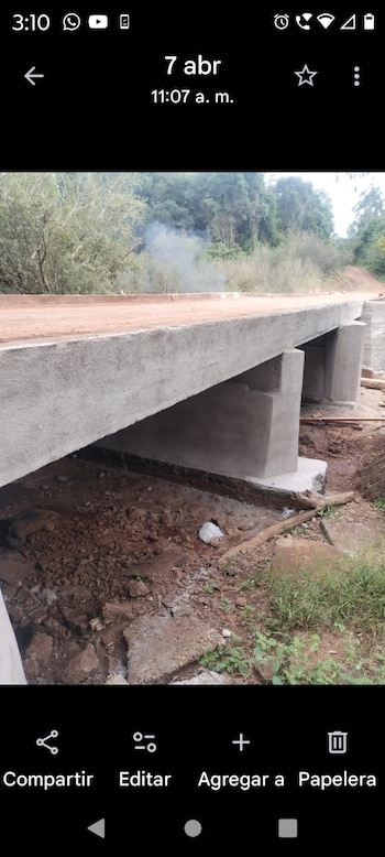 Un puente de concreto en construcción cruza un río de agua marrón y orillas fangosas, rodeado de frondosos árboles verdes bajo un cielo azul claro