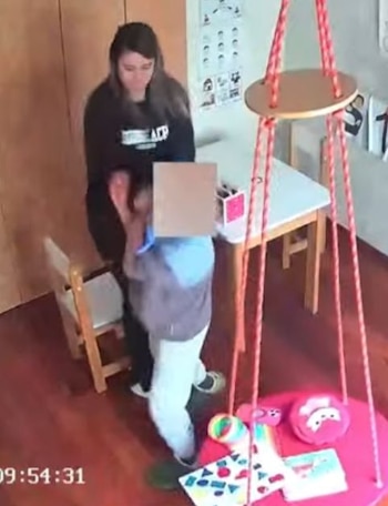 Una mujer de pie detrás de un niño con rostro censurado en una habitación con paredes de madera. Una mesa, una silla y una estructura con cuerdas y juguetes