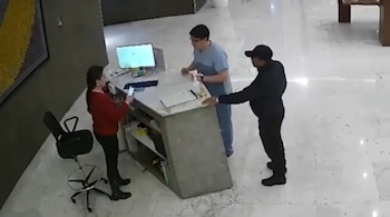 Una mujer con camiseta roja, dos hombres (uno con uniforme azul, otro con gorra negra), un mostrador, un monitor, un teléfono, una silla de oficina.