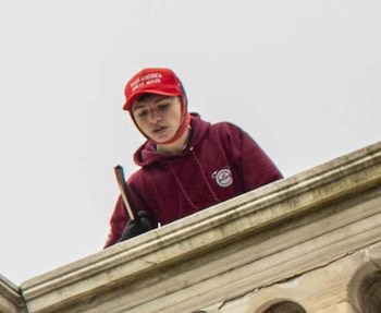 Un hombre joven, Elias Irizarry, con gorra roja 'MAGA' y sudadera granate, sostiene un objeto oscuro mientras mira hacia abajo desde un alféizar de piedra