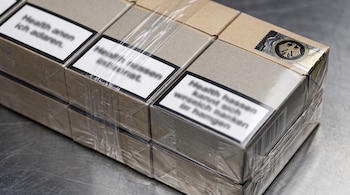 Primer plano de fajos de cigarrillos genéricos envueltos en plástico transparente, con etiquetas borrosas y un sello aduanero con águila mexicana estilizada.