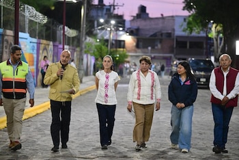 Claudia Sheinbaum y Delfina Gómez Álvarez, junto a otros cinco funcionarios, caminan por una calle adoquinada e iluminada al anochecer, con edificios al fondo