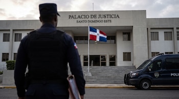 Agente de policía de espaldas con carpeta ante el Palacio de Justicia de Santo Domingo Este, la bandera dominicana y un vehículo policial.
