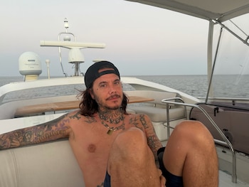 Sebastián Ortega con el torso descubierto y gorra negra sentado en la cubierta de un barco en el mar