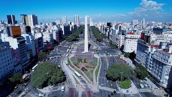 Vista aérea del Obelisco de Buenos Aires en el centro de la Avenida 9 de Julio, rodeado de tráfico y edificios urbanos bajo un cielo azul claro