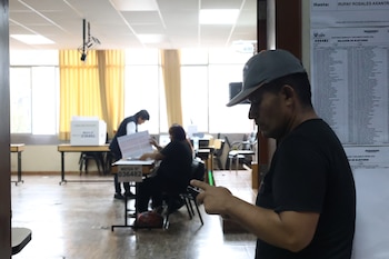 Un hombre con gorra y camisa oscura mira su teléfono en primer plano, mientras al fondo dos personas organizan una mesa de votación con una urna y documentos electorales en un salón