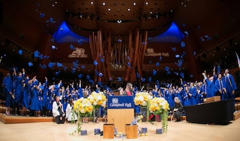 Estudiantes vestida con túnicas azules lanzan sus birretes al aire en un auditorio frente a un podio con el nombre Campbell Hall