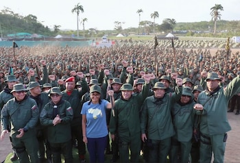 Multitud de personas con uniformes verdes y sombreros tipo pescador. Muchos puños levantados. Mujer con camiseta azul y mapa de Venezuela. Palmeras y tiendas de campaña