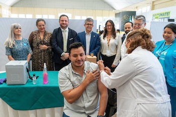 Un hombre recibe una inyección en el brazo. Una mujer con bata blanca aplica una jeringa. Varias personas de pie detrás. Mesa con hielera, botellas y cartel con texto