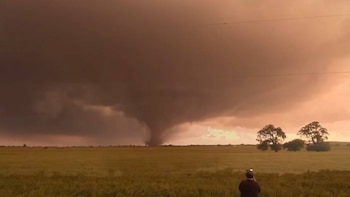 Un tornado grande y oscuro desciende de nubes de tormenta anaranjadas sobre un campo verde. En primer plano, una persona de espaldas observa