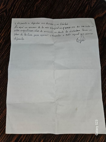 Una hoja de papel blanco con pliegues y texto manuscrito en tinta oscura, apoyada sobre una mesa de madera oscura
