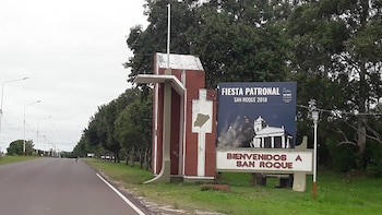 Entrada a localidad con señal de bienvenida a San Roque y cartel azul de Fiesta Patronal 2018. Carretera asfaltada, farolas, árboles verdes y cielo nublado