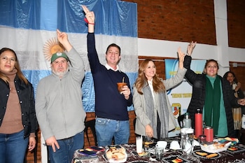 Cinco personas posan con los brazos levantados detrás de una mesa con vasos y comida; al fondo, una bandera argentina y una pared de ladrillos