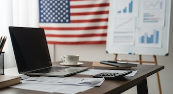 Mesa de estudio con laptop, papeles, calculadora y taza de café. Al fondo, bandera de Estados Unidos y pizarra con gráficos financieros borrosos.