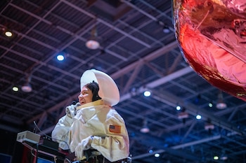 Persona en traje de astronauta blanco con bandera de EE. UU., sonríe con micrófono y panel de control. Luces y objeto rojo reflectante en el fondo
