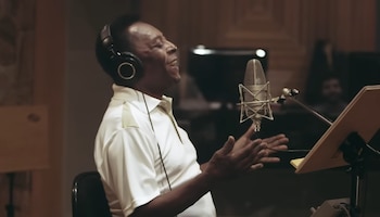 Hombre con camisa blanca y auriculares canta en un micrófono de estudio. Gesticula con las manos. Detrás hay un atril con una hoja de papel